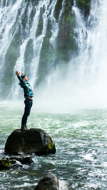 Man standing on large rock next to a waterfall with hands in the air